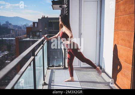 Beautiful Asian smiling fit woman doing yoga on sunny balcony in sport suit. Young happy lady on terrace at summertime. Big city view. Sports exercises and gymnastics, healthy way of life concept Stock Photo
