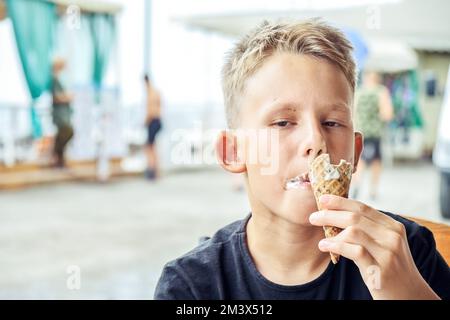 Junior schoolboy enjoys eating ice cream and spending summer holidays at sea resort. Blond-haired boy eats ice cream with satisfied expression Stock Photo