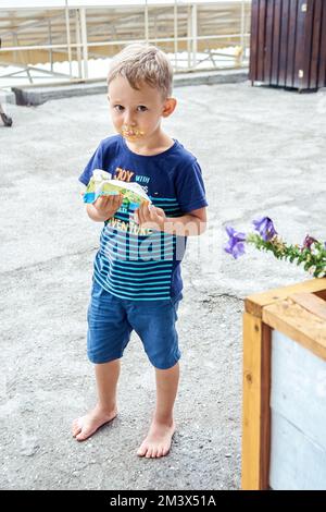 Preschooler boy gets dirty face eating favourite ice cream on sea resort street. Blond-haired guy enjoys spending summer holidays and resting Stock Photo