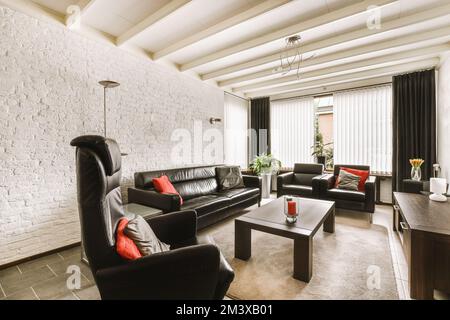 a living room with black leather couches and coffee table in front of white brick wall behind the sofa is dark wood Stock Photo