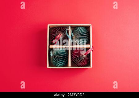 Trendy decorative paper Christmas tree baubles in a cardboard on red background. Top view, flat lay. Stock Photo