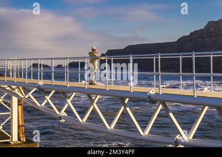 Solitary Lady stood on the East Pier bridge, wave-watching and admiring the scenery in Whitby (cliffs and Saltwick Nab) Stock Photo