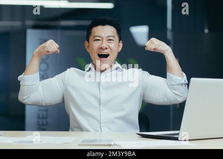 Portrait of a young happy Asian man at the desk in the office celebrating success, victory. He looks at the camera, shows a gesture of strength with his hands. Stock Photo