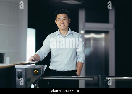 Young handsome Asian male worker enters office through electronic turnstile, automatic security system, gate. Encloses his electronic access card. He looks at the camera, smiles. Stock Photo