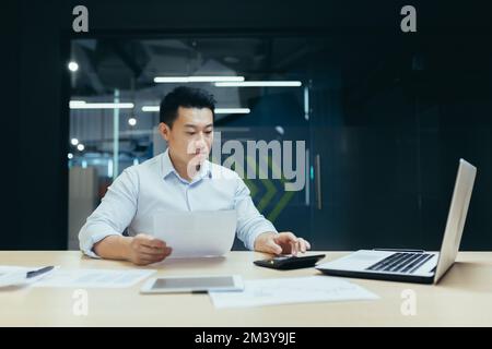 Portrait of a young male Asian accountant. He sits at the desk in the office, works with documents, accounts on a calculator and a laptop Stock Photo