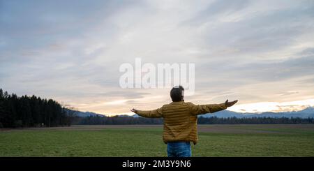 Young man in ochre colored jacket mindfully standing in beautiful green nature under cloudy evening sky with his arms wide open. Stock Photo