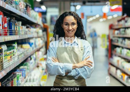 Portrait of Hispanic woman seller in supermarket, female worker in apron with curly hair and glasses smiling and looking at camera, saleswoman among goods with household chemicals and shelves. Stock Photo