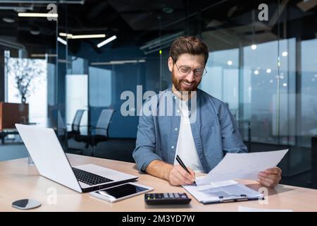 Mature businessman in shirt doing paperwork, man working with documents, contracts and bills sitting at table using laptop at work, financier accountant with beard and glasses. Stock Photo