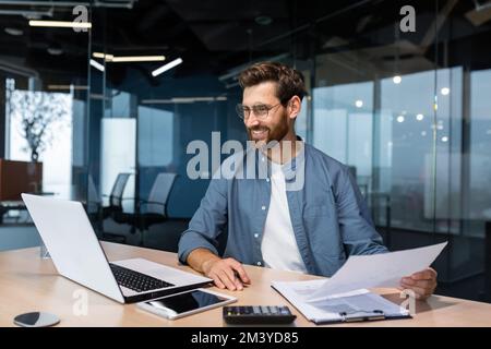 Mature businessman in shirt doing paperwork, man working with documents, contracts and bills sitting at table using laptop at work, financier accountant with beard and glasses. Stock Photo
