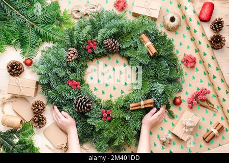 Child's hands holding handmade Christmas wreath on wrapping paper background, various accessories including fir tree branhes, pinecones and gifts, selective focus Stock Photo