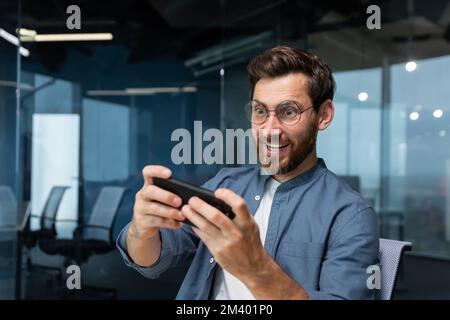 Mature businessman with beard is playing game on smartphone, man is having fun during break at workplace, businessman inside modern office. Stock Photo