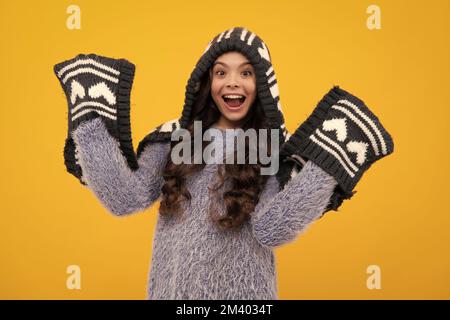 Amazed teenager. Warm hat with hood and scarf. Modern teen girl wearing sweater and knitted hat on isolated yellow background. Excited teen girl. Stock Photo