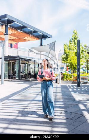 Confidence. Smiling latin university student woman walking and holding her books outside of the mall.  Stock Photo