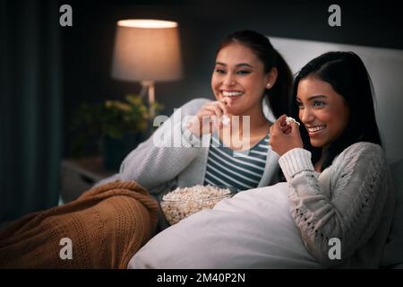 We never watch an episode alone. two young woman eating popcorn while sitting together on a bed at night. Stock Photo