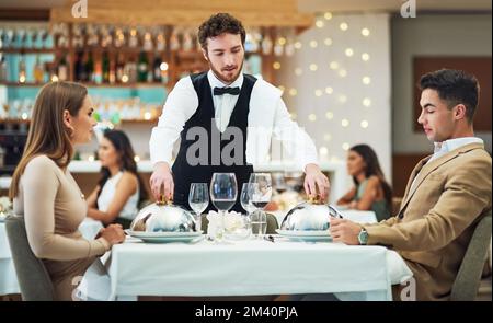 Allow me to serve you. a handsome young waiter serving a young couple in a restaurant. Stock Photo