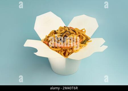 Udon noodles with fried chicken, seafood and vegetables in the cardboard box with wooden chopsticks on the bright blue background color. Stock Photo