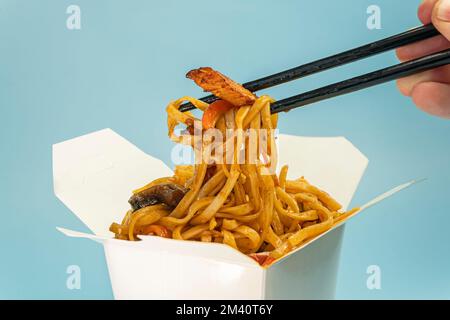 Udon stir-fry noodles with chicken meat and sesame in cardboard box on blue background close up. bright blue background color. Stock Photo