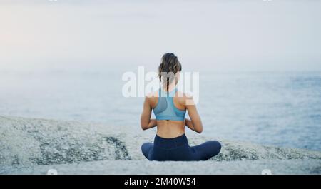 The perfect setting for perfect calm. Rearview shot of an unrecognizable woman sitting cross legged and meditating alone by the ocean during an Stock Photo