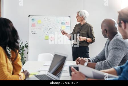 We all have to work together to achieve our target. a mature businesswoman discussing ideas on a whiteboard with her colleagues at work. Stock Photo