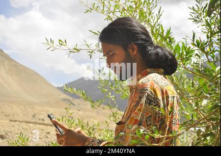 Side view of a young man with long hair and beard using phone with the background of mountains of Zanskar valley. Stock Photo