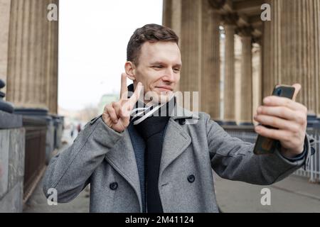 Handsome young man in coat walking on street holds mobile phone and shoots video or takes photo, takes a selfie on smartphone and communicates online via video communication Stock Photo