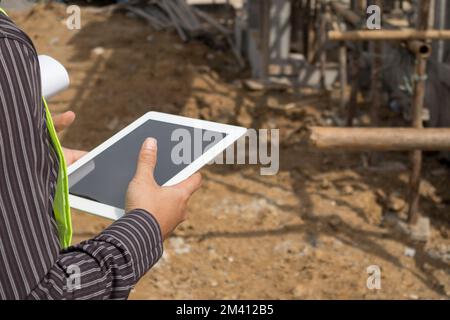 Asian business man construction manager and engineer worker in protective helmet hold blueprints paper on hand using tablet computer at building site Stock Photo