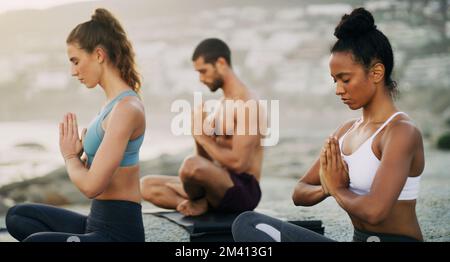 We are rooted but we flow. a group of young people sitting and meditating together while on the beach during an overcast day. Stock Photo