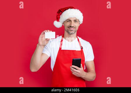 mature man in christmas santa hat and red apron hold smartphone and card. internet buy Stock Photo
