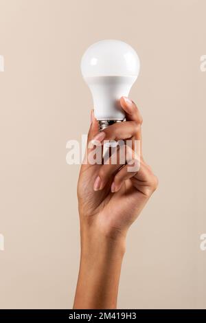 Anonymous black female demonstrating LED light bulb in raised arms while promoting energy saving ecology campaign against beige background Stock Photo