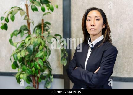 Self assured Asian female entrepreneur in suit crossing arms and looking at camera while standing near lush potted plant in office Stock Photo