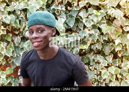 Glad African American male in t shirt and cap looking at camera with smile near green leaves in garden Stock Photo