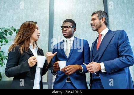 Low angle of Asian female entrepreneur in formal wear with mug of hot drink gesticulating and speaking with diverse male colleagues during coffee brea Stock Photo