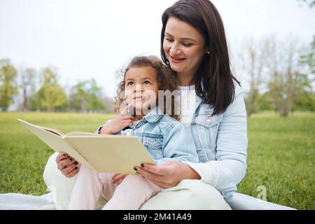 Children park, reading and mother with a girl and story book with happiness from creative development. Happy, relax and interracial family on a picnic Stock Photo