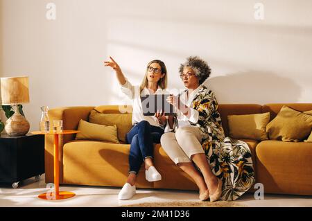 Creative interior designers discussing a new idea while sitting in an office lobby. Two businesswomen using a digital tablet while working together in Stock Photo