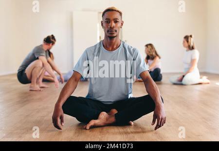Portrait of a male yogi looking at the camera while sitting in lotus position with a group of people in the background. Young black man attending a yo Stock Photo