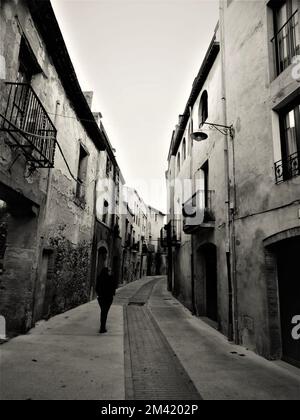 A vertical grayscale shot of a person walking down the narrow street surrounded by old buildings. Stock Photo