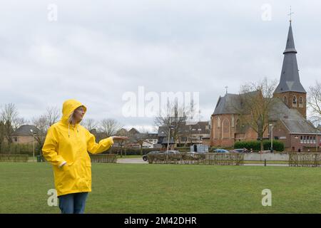 Waiting for the rain. Woman in a yellow raincoat on a grassy meadow against the backdrop of an old catholic church Stock Photo