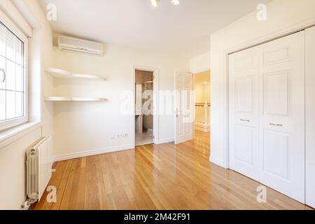 Empty room with fitted wardrobes with white lacquered doors, built-in shelves in one corner, en-suite marble bathroom and pine wood flooring Stock Photo