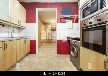 Kitchen furnished with light wood furniture, red wall and white tiles and light brown stoneware floors Stock Photo