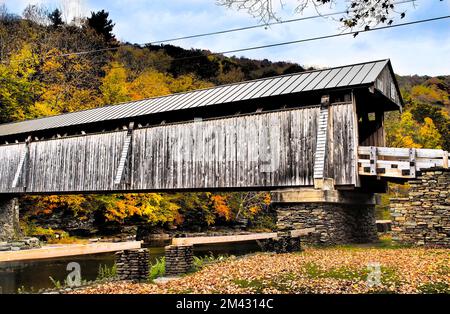 Rural covered bridge over calm river, vibrant Autumn leaf colors in the forested hills, and blue sky. With filters for retro postcard effect. Stock Photo