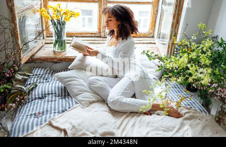 Pretty romantic african american girl, reading book sitting on windowsill, suraunded with flowers. Beautiful Afro woman happily reading a novel in coz Stock Photo