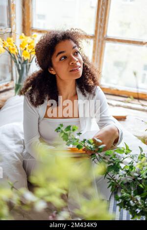 Pretty romantic african american girl, reading book sitting on windowsill, suraunded with flowers. Beautiful Afro woman happily reading a novel in coz Stock Photo