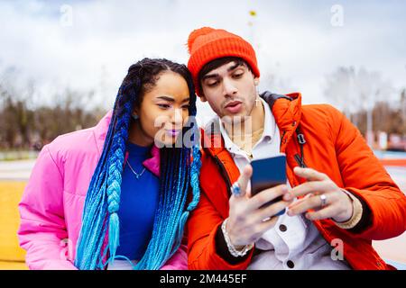 Multiracial young couple of lovers dating outdoors in winter using social media app on smartphone, wearing winter jackets and having fun - Multiethnic Stock Photo