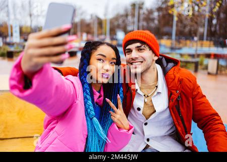 Multiracial young couple of lovers dating outdoors in winter using social media app on smartphone, wearing winter jackets and having fun - Multiethnic Stock Photo