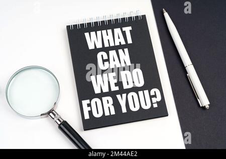 Business and economy concept. On a black and white surface lies a magnifying glass, a pen and a notepad with the inscription - WHAT CAN WE DO FOR YOU Stock Photo