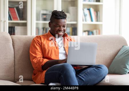 Happy African American Guy Using Laptop Computer Sitting On Couch Stock Photo