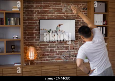 Sporty middle aged man working out at home, watching sport channel and fitness workout program on TV, back view Stock Photo