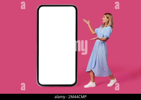 Smiling shocked mature caucasian lady blonde in dress shows with hands on big smartphone with blank screen Stock Photo
