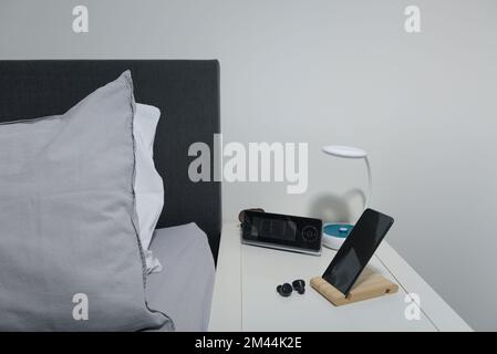 Actual for the bedroom. smartphone on the bedside table with lamp, headset, clock and bed. Stock Photo