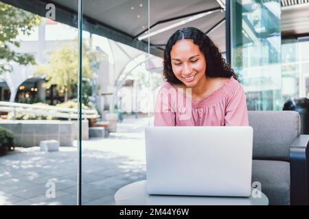 Happy young latin student woman using a laptop and sitting outside a cafe in her free time Stock Photo